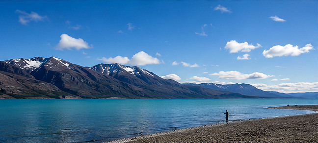 The turquoise water of Jurassic Lake below snow-dusted Patagonian mountains, an angler wading the shoreline